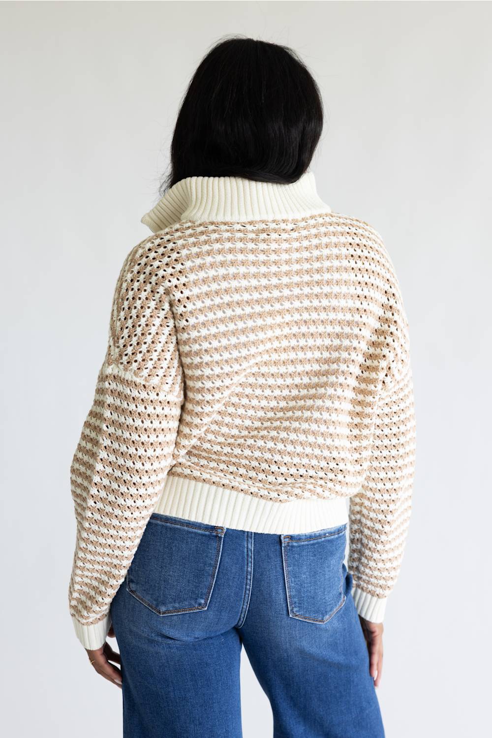 A person shown from the back wearing a cream and beige open-knit cropped sweater with a ribbed turtleneck and hem, paired with blue jeans, against a plain light gray studio background.