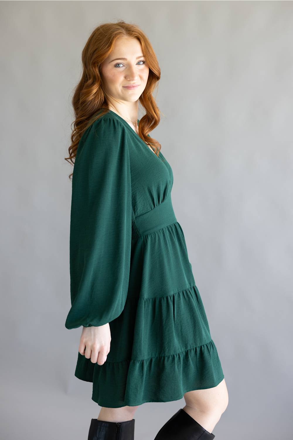 A woman modeling a dark green tiered mini dress with long balloon sleeves and black knee-high boots, standing against a neutral gray studio backdrop.