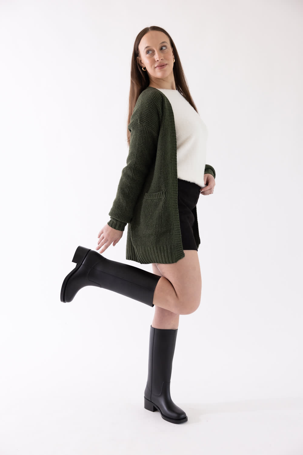 A woman modeling a dark olive green knit cardigan over a white top and black shorts, paired with black knee-high boots, against a plain white studio background.