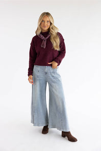 A woman modeling a dark burgundy knit sweater with a matching neck scarf, paired with wide-leg light blue jeans and brown ankle boots, against a plain white studio background.