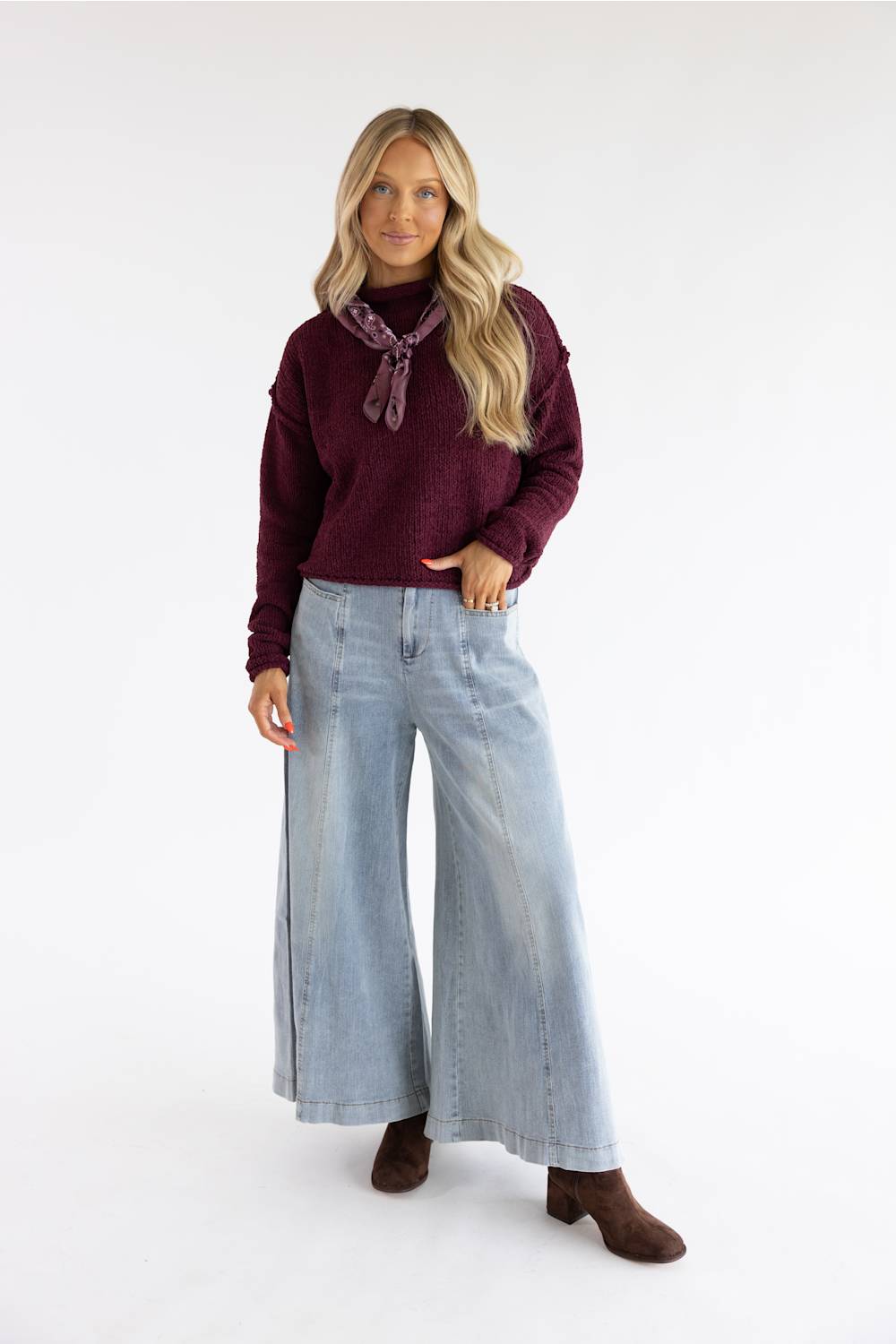 A woman modeling a dark burgundy knit sweater with a matching neck scarf, paired with wide-leg light blue jeans and brown ankle boots, against a plain white studio background.