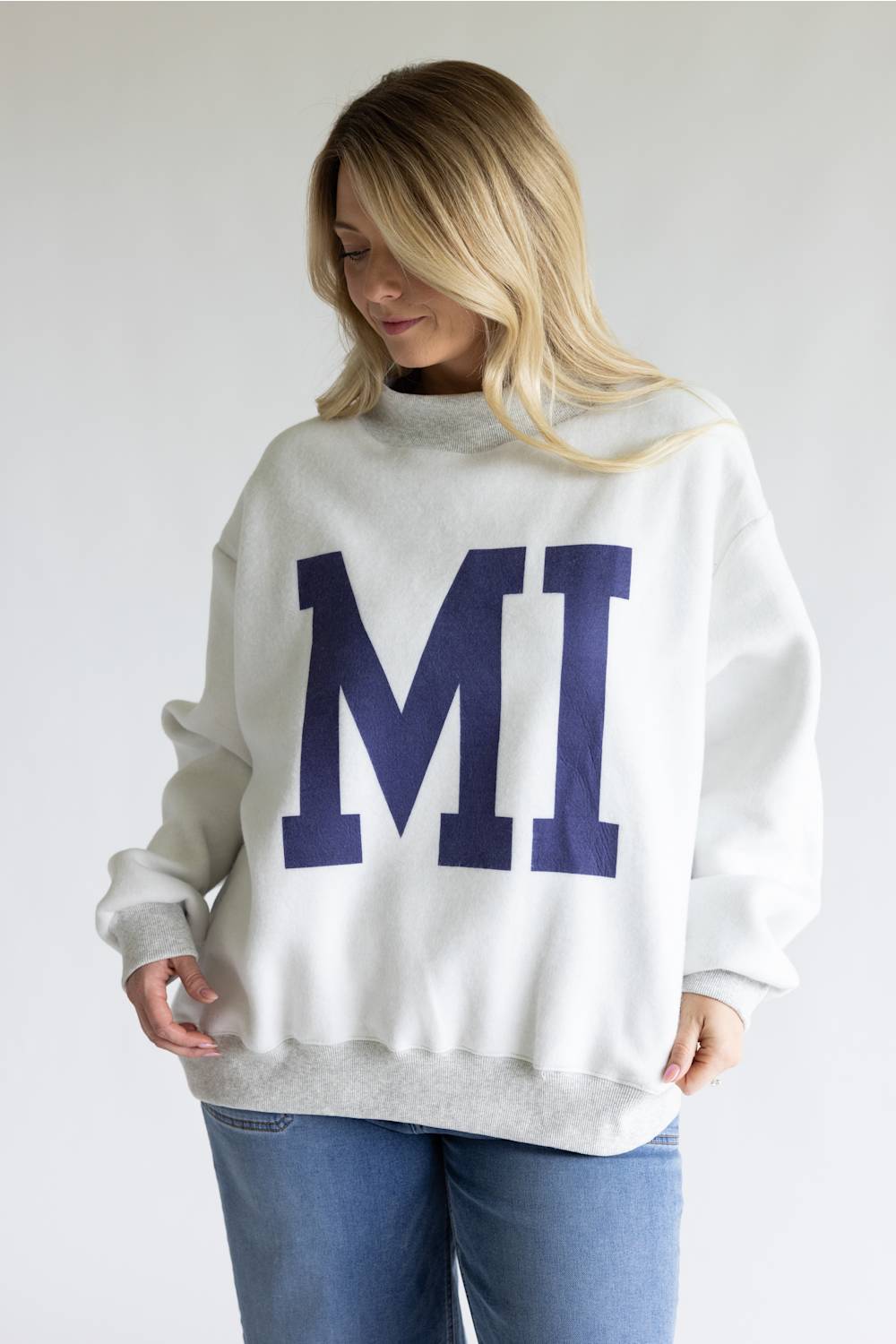 A woman wearing an oversized white crewneck sweatshirt with a large navy blue 'MI' graphic on the front, paired with blue jeans, against a plain light gray background.