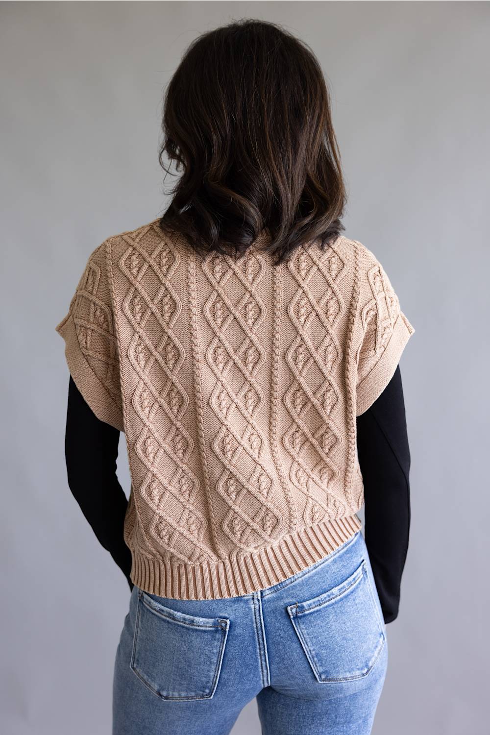A woman viewed from behind wearing a beige cable-knit cropped sweater vest over a black long-sleeve shirt and blue jeans, against a plain gray studio background.