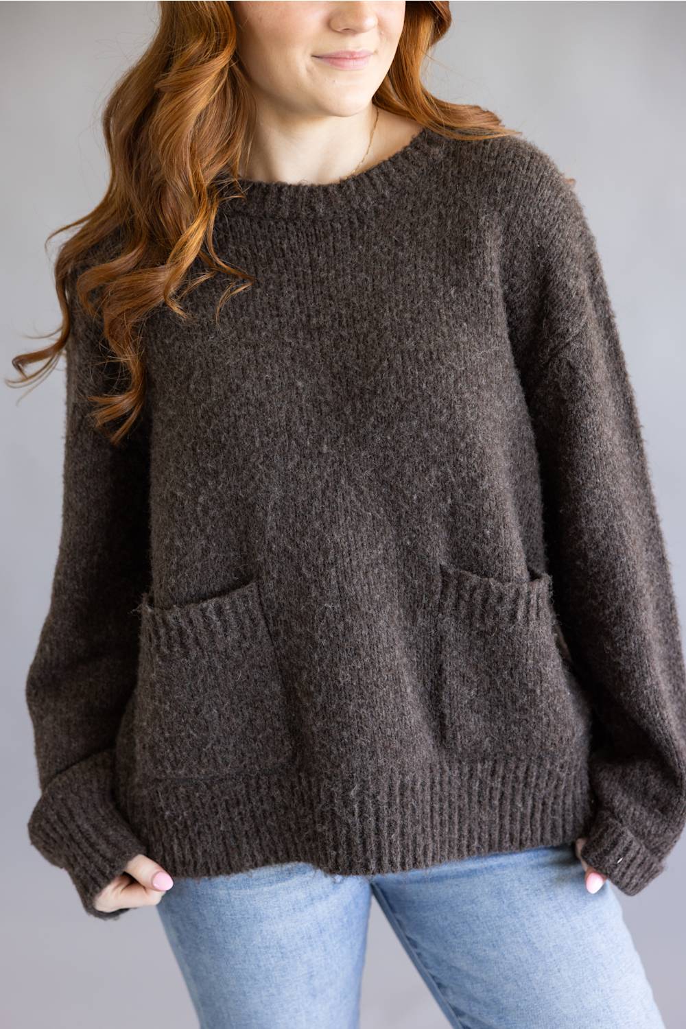  oversized, chunky knit brown sweater with front pockets and light blue jeans, standing against a plain gray studio background
