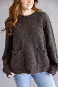  oversized, chunky knit brown sweater with front pockets and light blue jeans, standing against a plain gray studio background