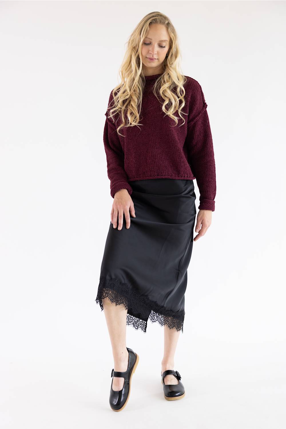 A woman modeling a dark burgundy knit sweater and a black midi skirt with a lace hem, paired with black Mary Jane shoes, against a plain white studio background.