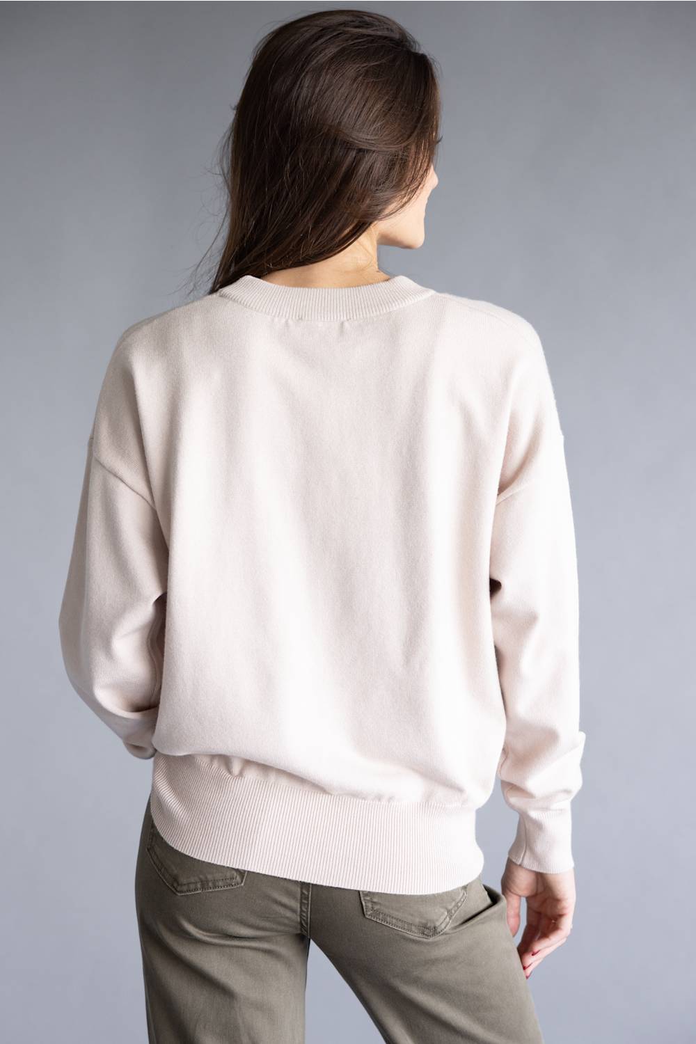 A woman viewed from the back wearing a light cream-colored crewneck sweatshirt and olive green pants, set against a solid gray studio background.