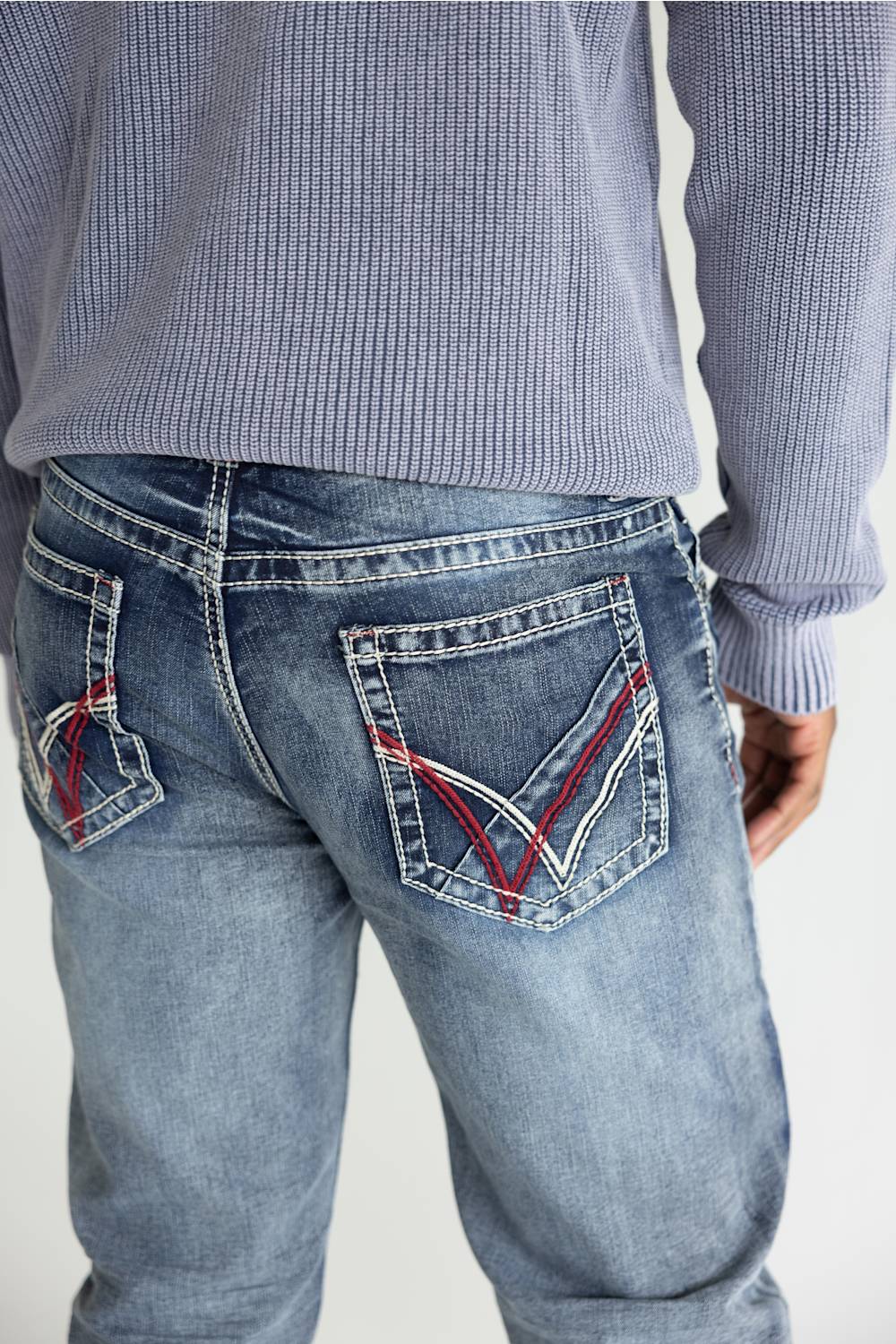 Close-up back view of a person wearing light-wash blue jeans with distinctive red and white contrast stitching on the back pockets, paired with a ribbed long-sleeve top against a plain light background.