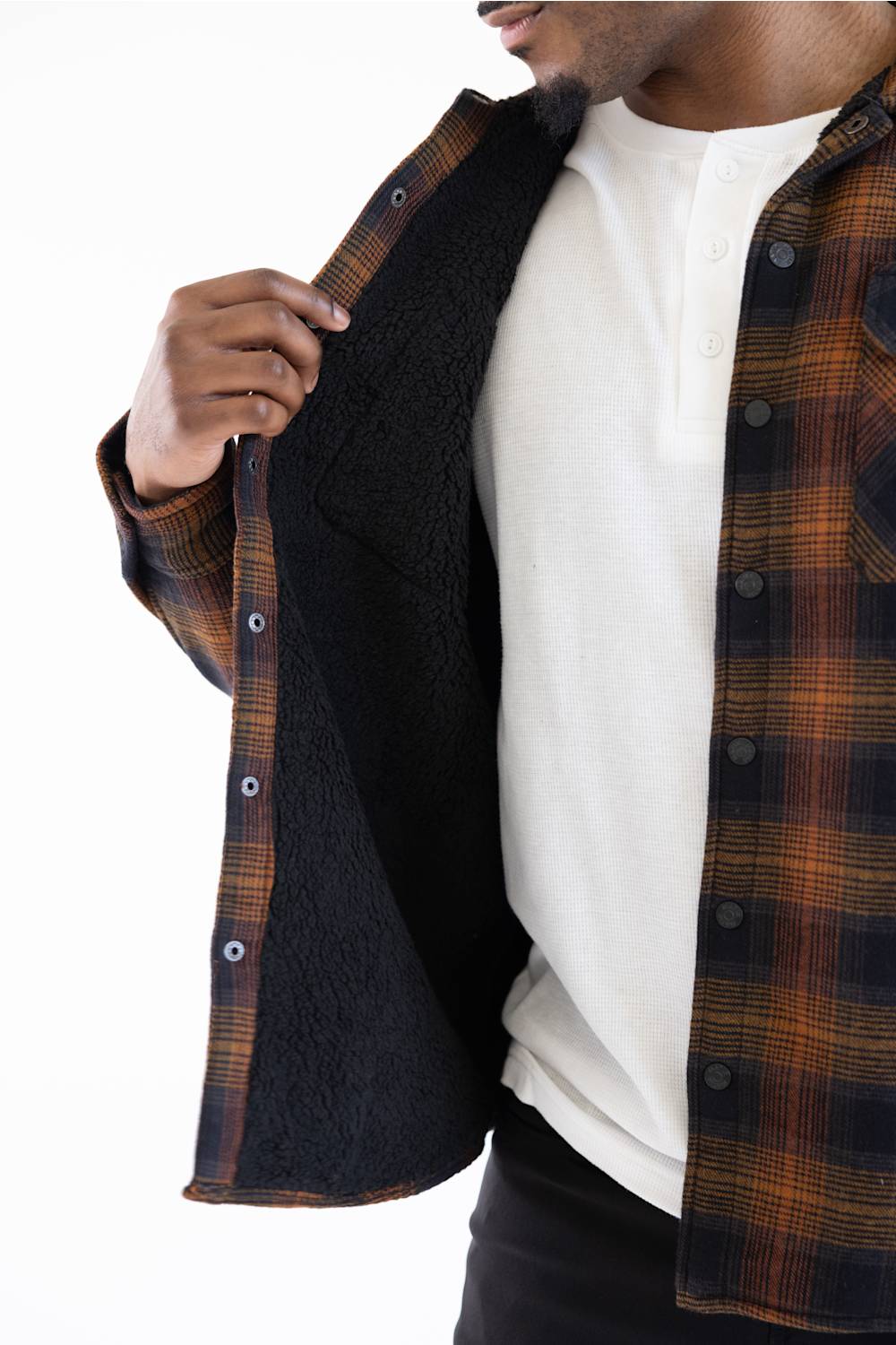 A close-up of a man wearing a brown and black plaid flannel shirt with a black sherpa lining, layered over a white henley shirt, against a white background.