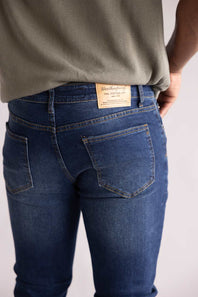 Close-up of the back pockets of a person wearing blue jeans with a visible brand label on a plain background. The blue jeans are named Weatherproof Vintage Slim Jeans for Men in Silt Blue.