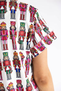 A close-up of a person wearing a white short-sleeve top with a colorful all-over print of nutcracker figures. The print features multiple nutcrackers in red, green, pink, blue, and gold uniforms.