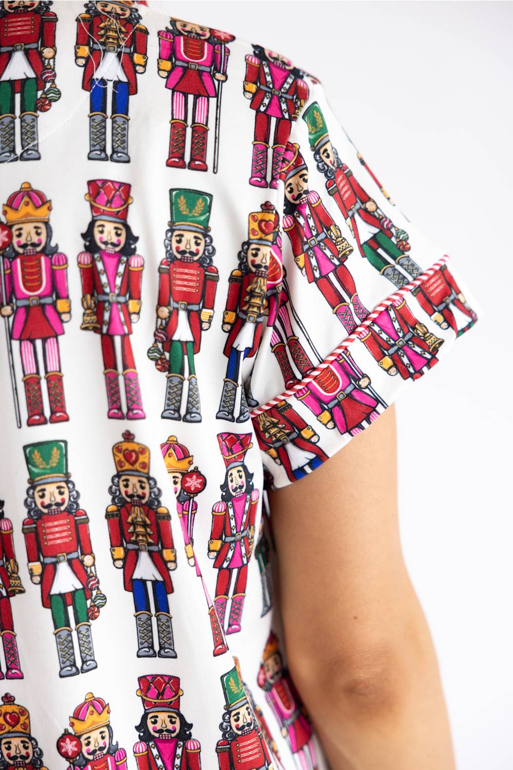 A close-up of a person wearing a white short-sleeve top with a colorful all-over print of nutcracker figures. The print features multiple nutcrackers in red, green, pink, blue, and gold uniforms.