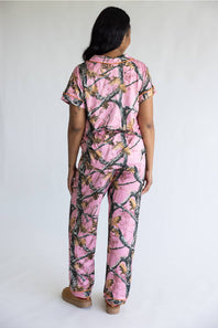 A woman viewed from behind wearing a pink camouflage-print short-sleeve top and matching pants, standing against a plain light gray studio background.