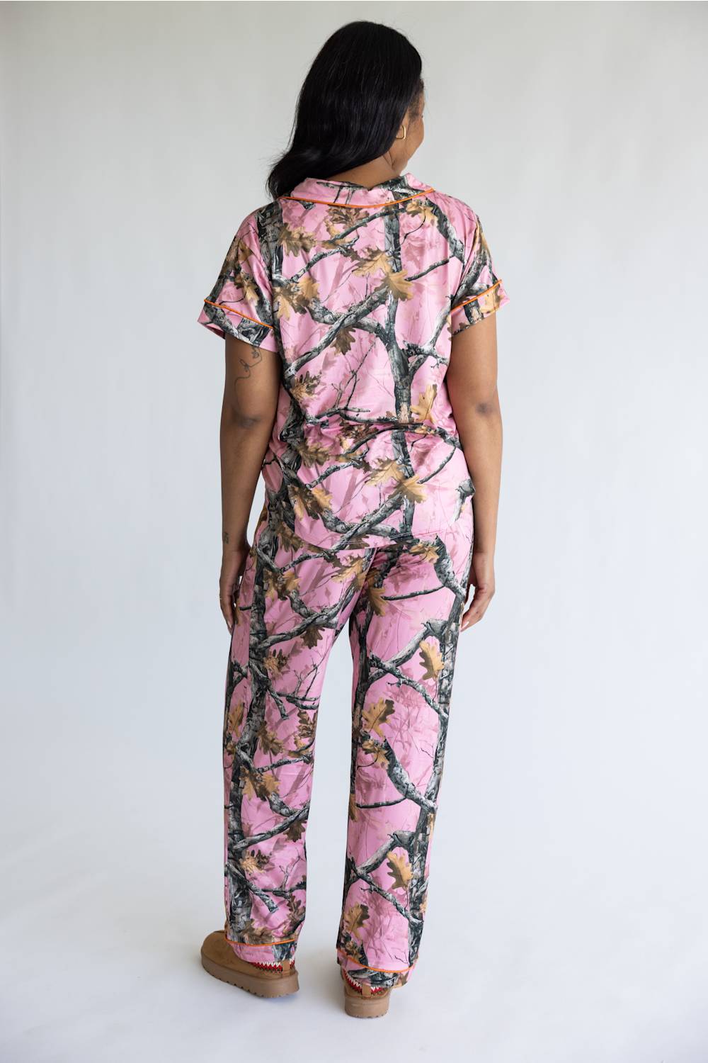 A woman viewed from behind wearing a pink camouflage-print short-sleeve top and matching pants, standing against a plain light gray studio background.