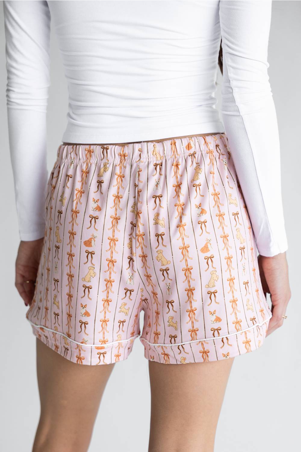 A person wearing light pink shorts with an allover print of bows and dogs  in orange, yellow, and brown, paired with a white long-sleeve top against a light gray studio background.