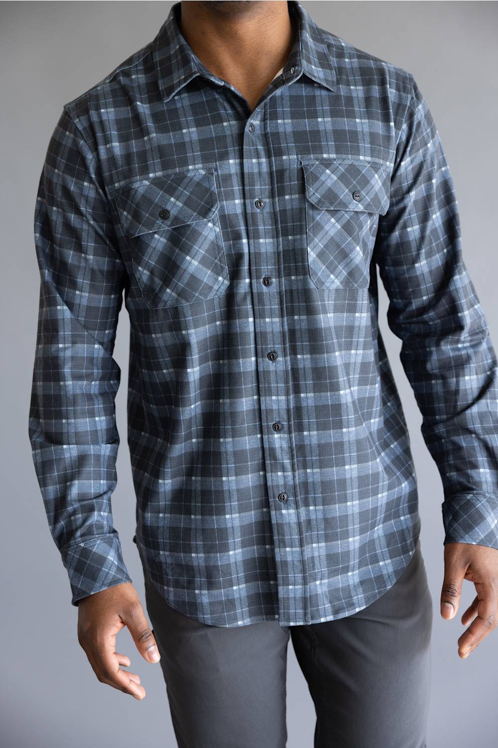 A man wearing a long-sleeve button-up plaid shirt in shades of blue, gray, and white, paired with dark gray pants, against a plain gray background.