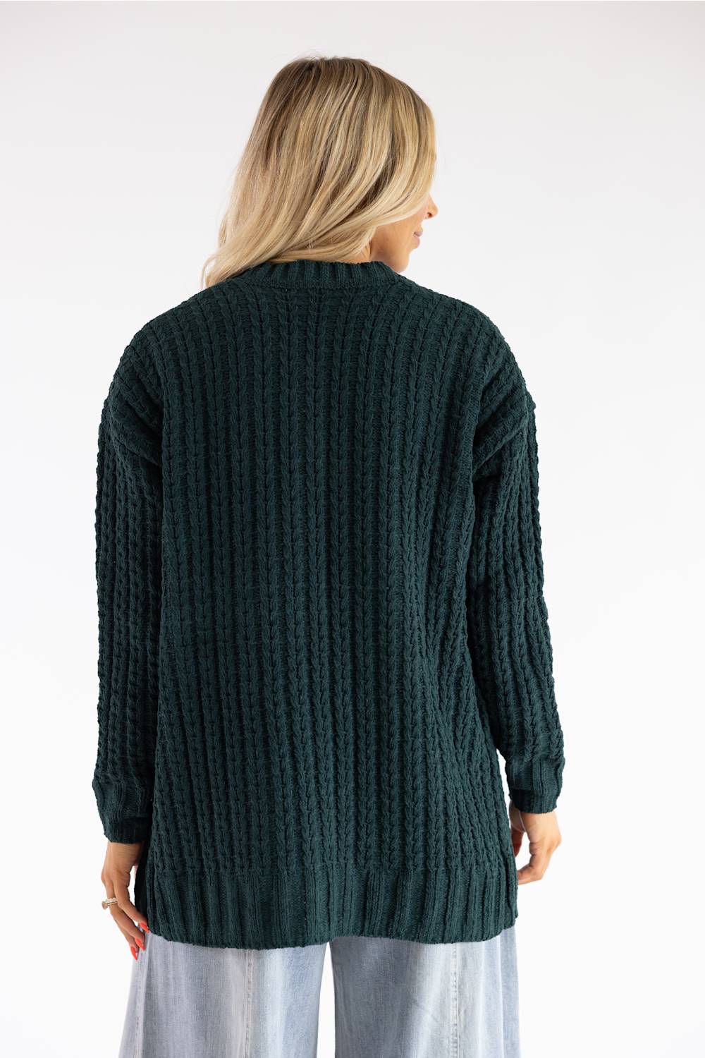 A woman viewed from the back wearing a dark forest green cable-knit sweater and light blue jeans against a plain white studio background.