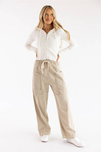 Three-quarter view of a model wearing a Pink Rose blizzard white cable-knit full-zip sweater hoodie styled with beige drawstring utility cargo pants against a plain backdrop.
