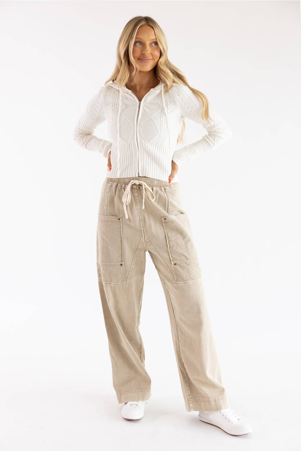 Three-quarter view of a model wearing a Pink Rose blizzard white cable-knit full-zip sweater hoodie styled with beige drawstring utility cargo pants against a plain backdrop.
