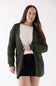 A woman modeling an olive green waffle-knit open-front cardigan over a cream sweater and black skirt, photographed against a plain white studio background.