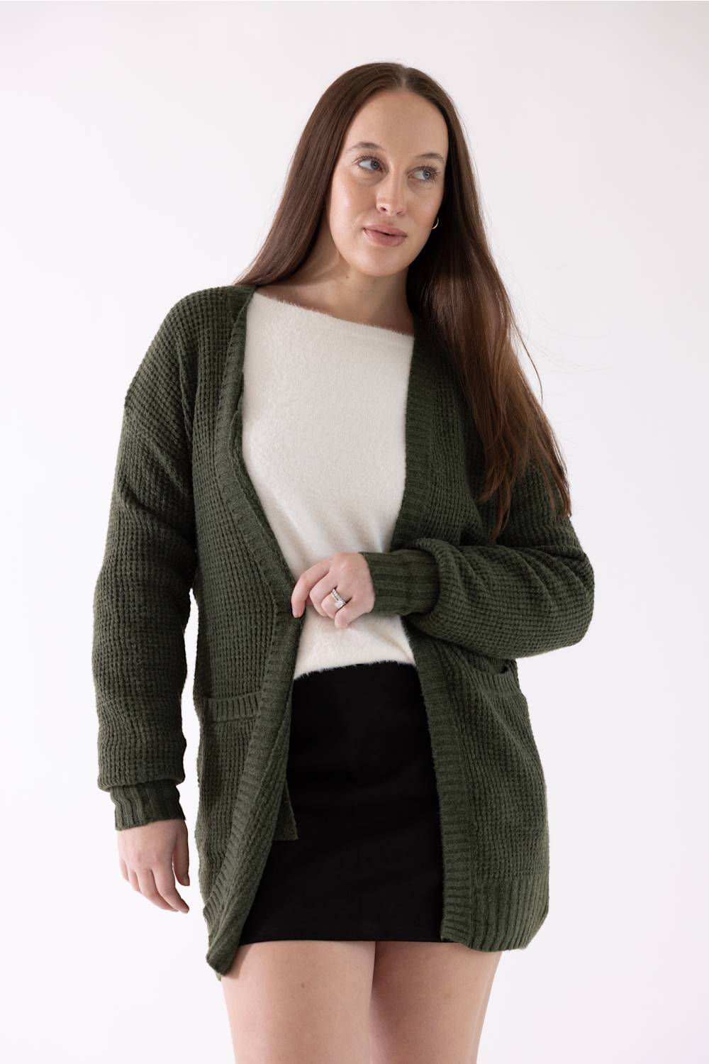 A woman modeling an olive green waffle-knit open-front cardigan over a cream sweater and black skirt, photographed against a plain white studio background.