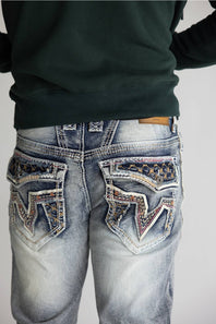 Close-up back view of a person wearing distressed light-wash blue jeans with heavily embellished decorative pocket patches and a dark green sweatshirt against a plain light gray background.