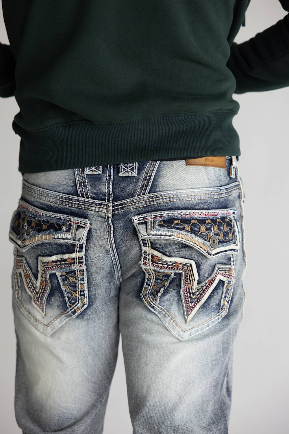 Close-up back view of a person wearing distressed light-wash blue jeans with heavily embellished decorative pocket patches and a dark green sweatshirt against a plain light gray background.
