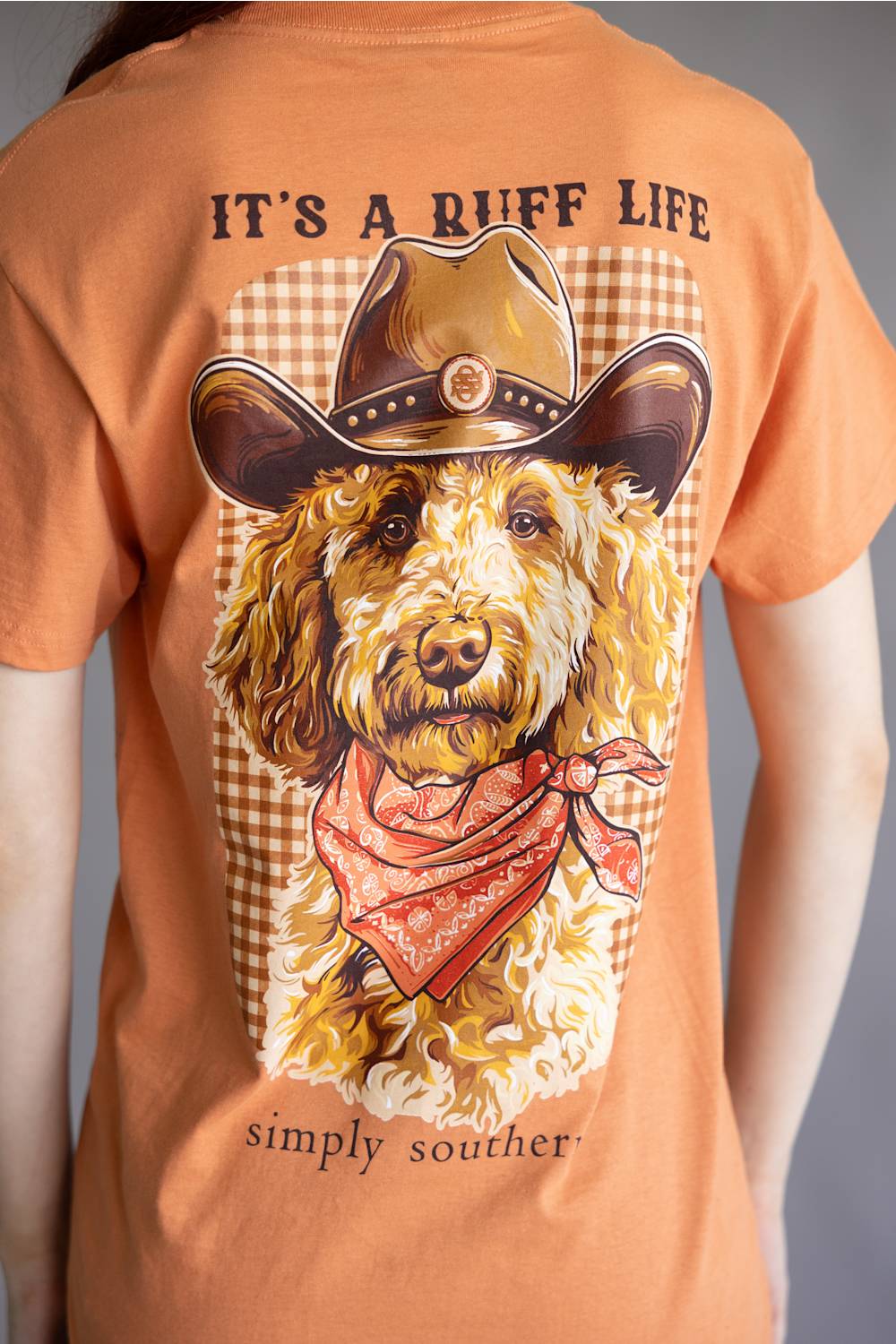 A close-up of a person wearing a peach-colored graphic t-shirt. The back of the shirt features a large illustration of a curly-haired dog wearing a cowboy hat and bandana, with the text 'IT'S A RUFF LIFE' above and 'simply souther.