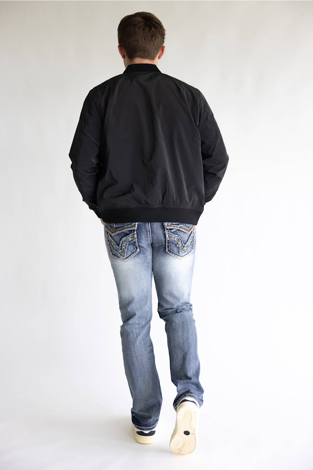A man viewed from behind wearing a black bomber jacket, light blue jeans, and white sneakers, standing against a plain white studio background.