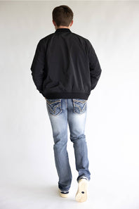 A man viewed from behind wearing a black bomber jacket, light blue jeans, and white sneakers, standing against a plain white studio background.