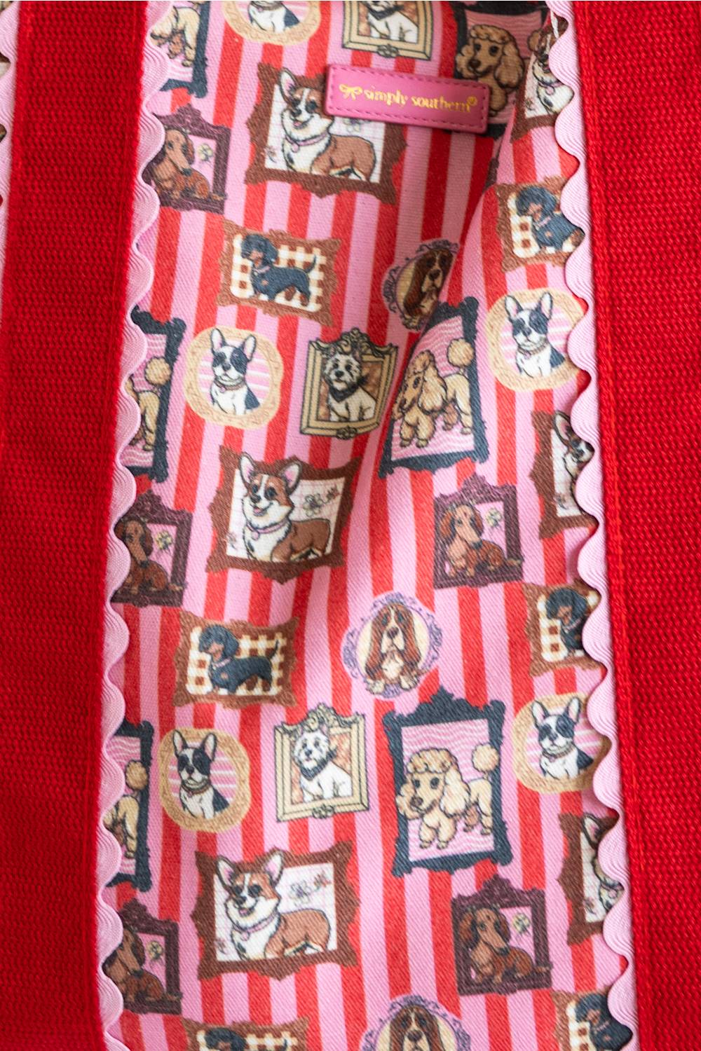 A close-up of a pink striped fabric accessory with scalloped edges, featuring a repeating pattern of dog portraits in picture frames. The fabric is laid over a red textured background, and a pink label at the top reads 'Simply Southern'.