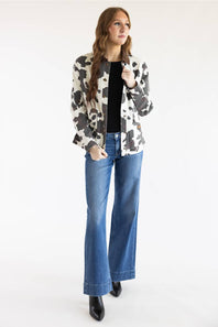 A woman modeling a black and white cow-print blazer over a black top and blue wide-leg jeans, standing against a plain white studio background.