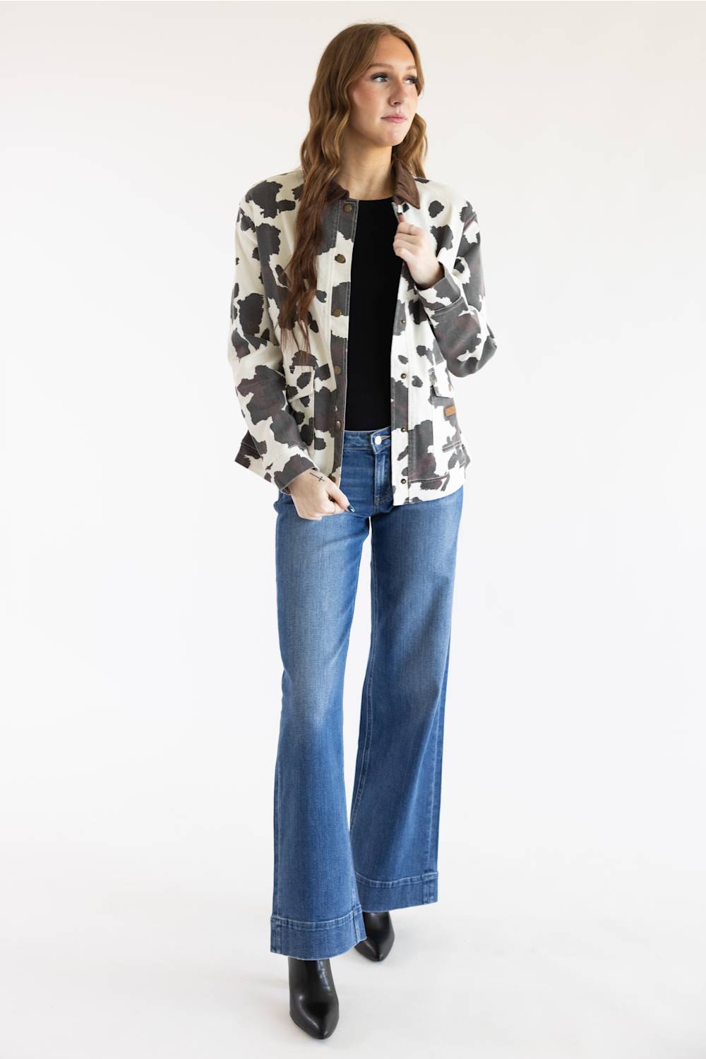A woman modeling a black and white cow-print blazer over a black top and blue wide-leg jeans, standing against a plain white studio background.