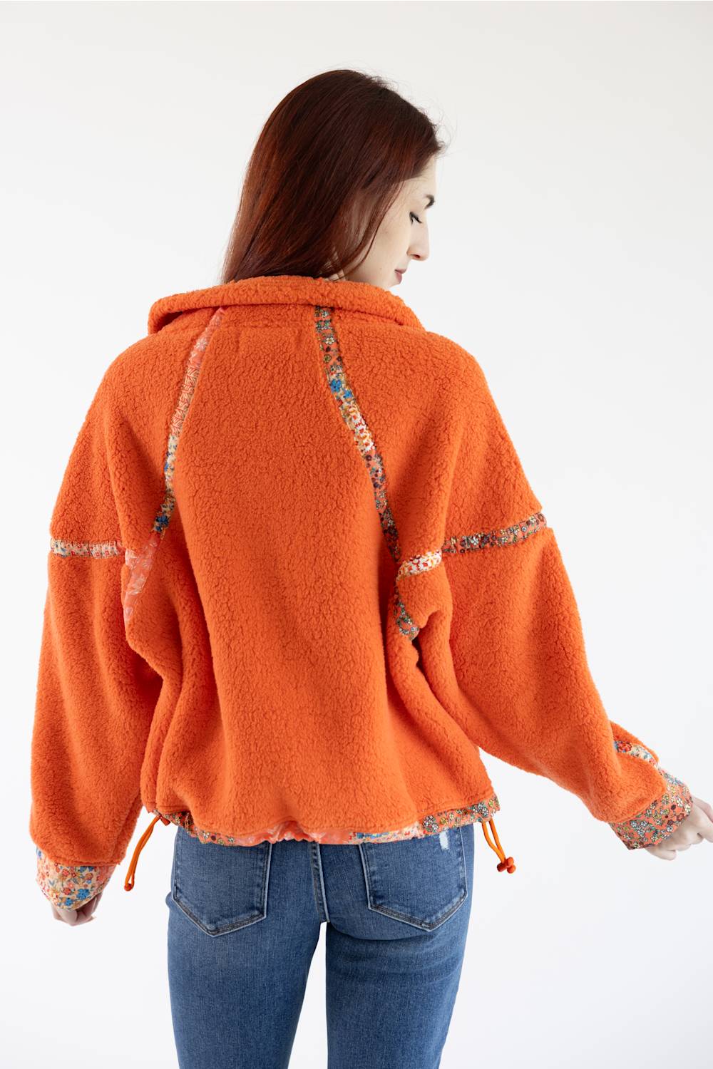 A woman shown from the back wearing a bright orange fuzzy cropped jacket with patterned trim details and blue jeans, against a plain white background.