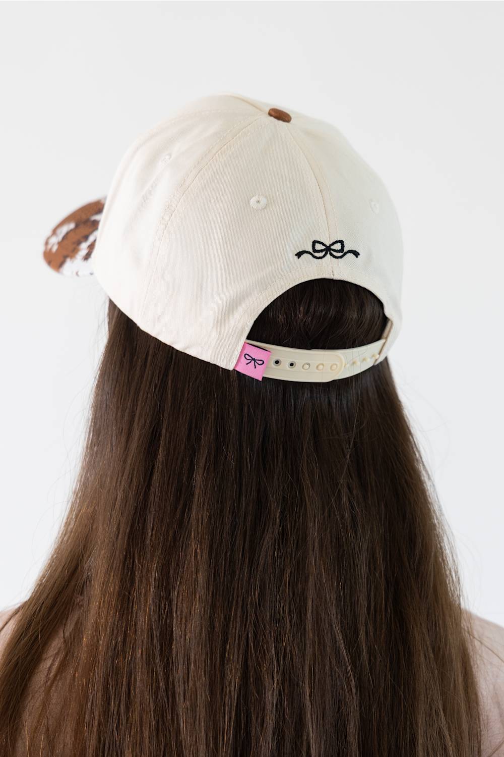A person viewed from behind wearing a cream-colored baseball cap with a small black bow embroidery on the back and a pink brand tag on the side. The person has long, straight brown hair.