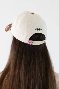 A person viewed from behind wearing a cream-colored baseball cap with a small black bow embroidery on the back and a pink brand tag on the side. The person has long, straight brown hair.