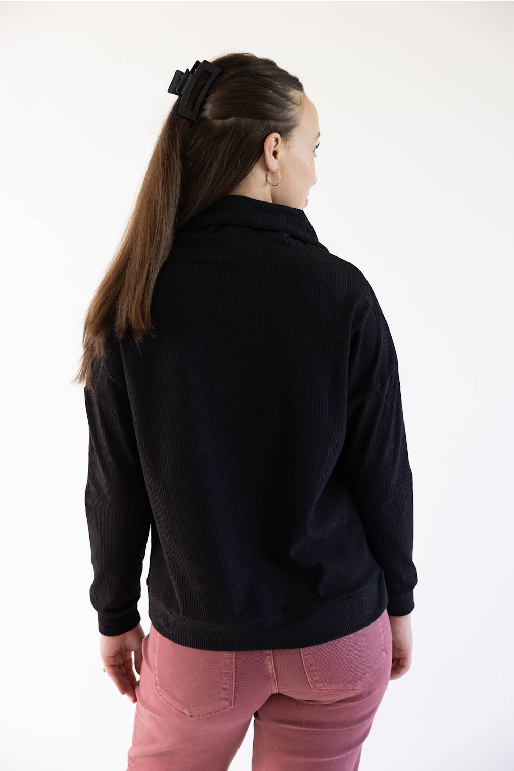 A woman shown from the back wearing a black long-sleeve collared top and pink pants, standing against a plain white background.