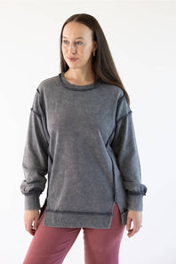 A woman modeling a dark gray mineral-wash crewneck sweatshirt with long sleeves and side slits, paired with dusty rose pants, against a plain white studio background.