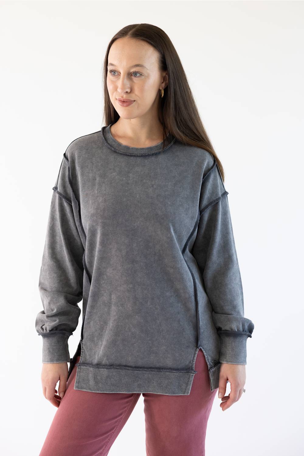 A woman modeling a dark gray mineral-wash crewneck sweatshirt with long sleeves and side slits, paired with dusty rose pants, against a plain white studio background.