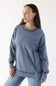 A woman wearing a washed slate blue crewneck sweatshirt and matching blue jeans, photographed against a clean white studio background.