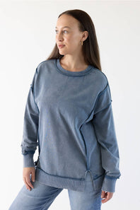 A woman wearing a washed slate blue crewneck sweatshirt and matching blue jeans, photographed against a clean white studio background.