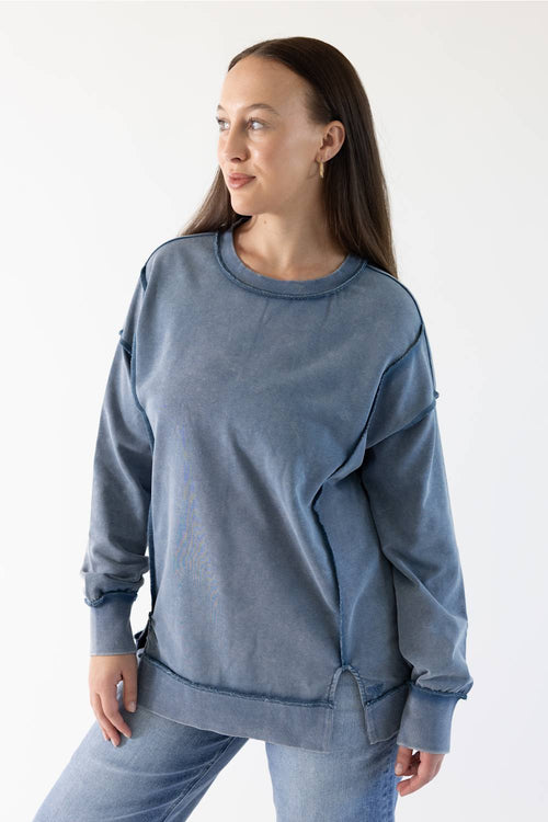 A woman wearing a washed slate blue crewneck sweatshirt and matching blue jeans, photographed against a clean white studio background.