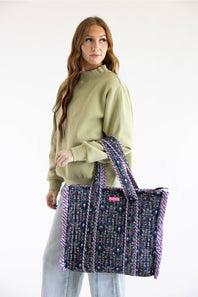 A woman wearing a sage green sweatshirt and light blue jeans holds a large woven tote bag with purple and multicolor patterned handles and trim against a plain white studio background.