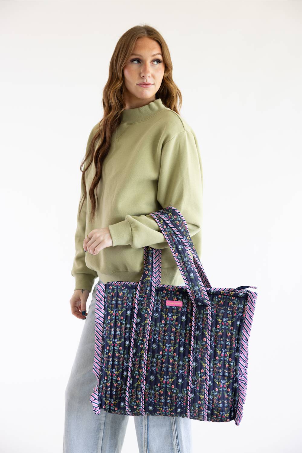 A woman wearing a sage green sweatshirt and light blue jeans holds a large woven tote bag with purple and multicolor patterned handles and trim against a plain white studio background.