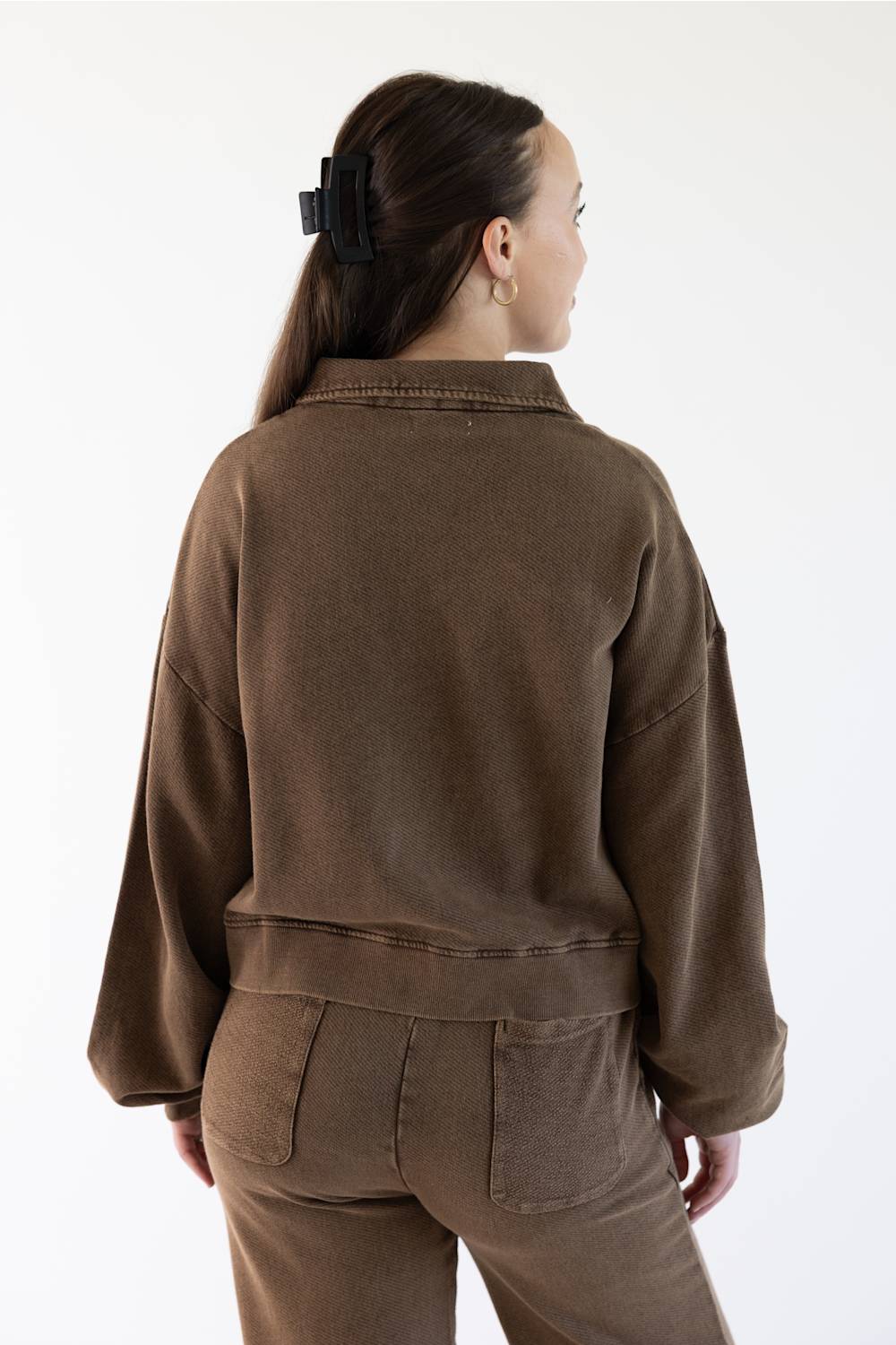 A woman shown from the back wearing a brown cropped sweatshirt and matching brown pants against a plain white studio background.