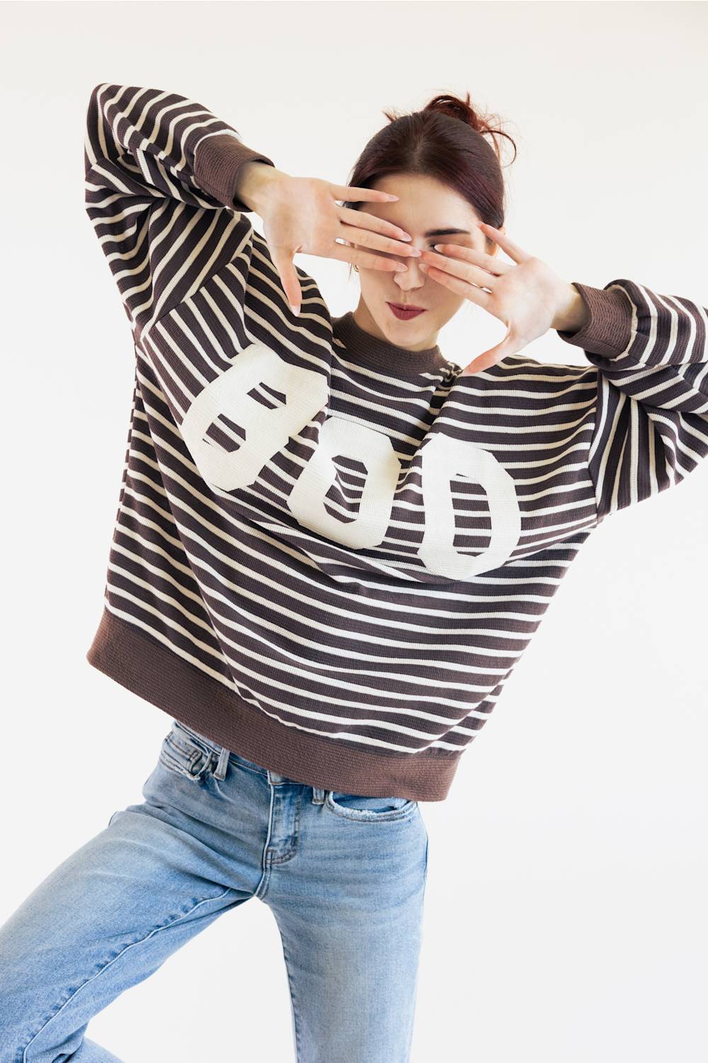 A woman posing in a brown and white horizontally striped sweatshirt with a large white BOO graphic on the front, paired with light blue jeans, against a plain white background.