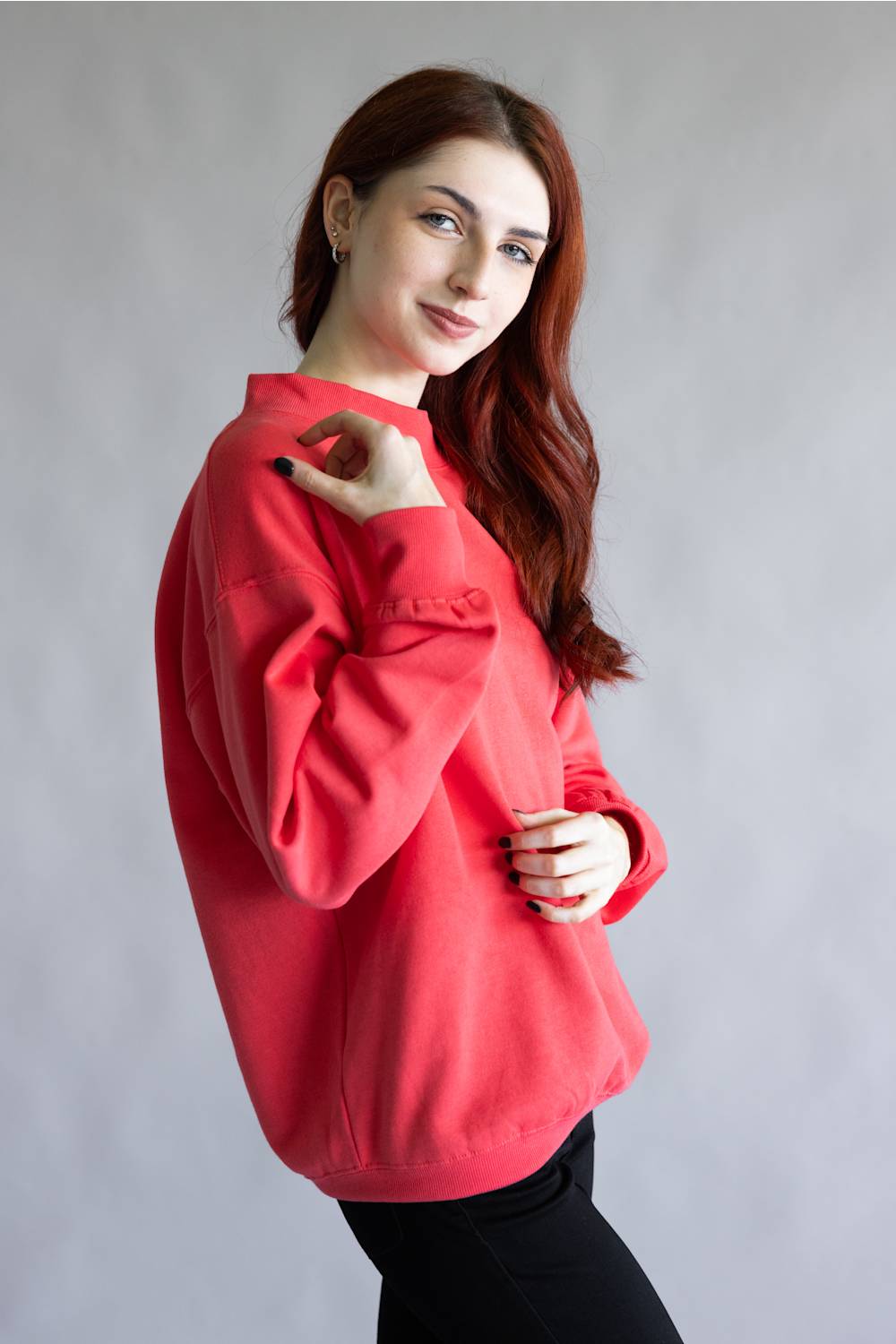 A woman with long wavy red hair wearing a bright red crewneck sweatshirt and black pants, posing against a neutral gray studio background.