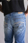 Close-up back view of a person wearing light blue denim jeans with decorative contrast stitching and a tan leather patch at the back waistband, paired with a black shirt against a light gray background.