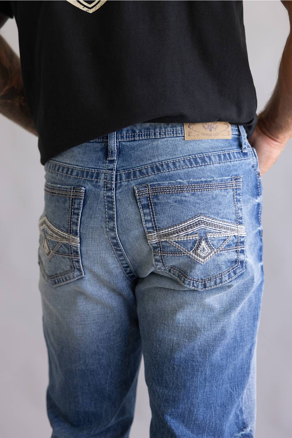 Close-up back view of a person wearing light blue denim jeans with decorative contrast stitching and a tan leather patch at the back waistband, paired with a black shirt against a light gray background.