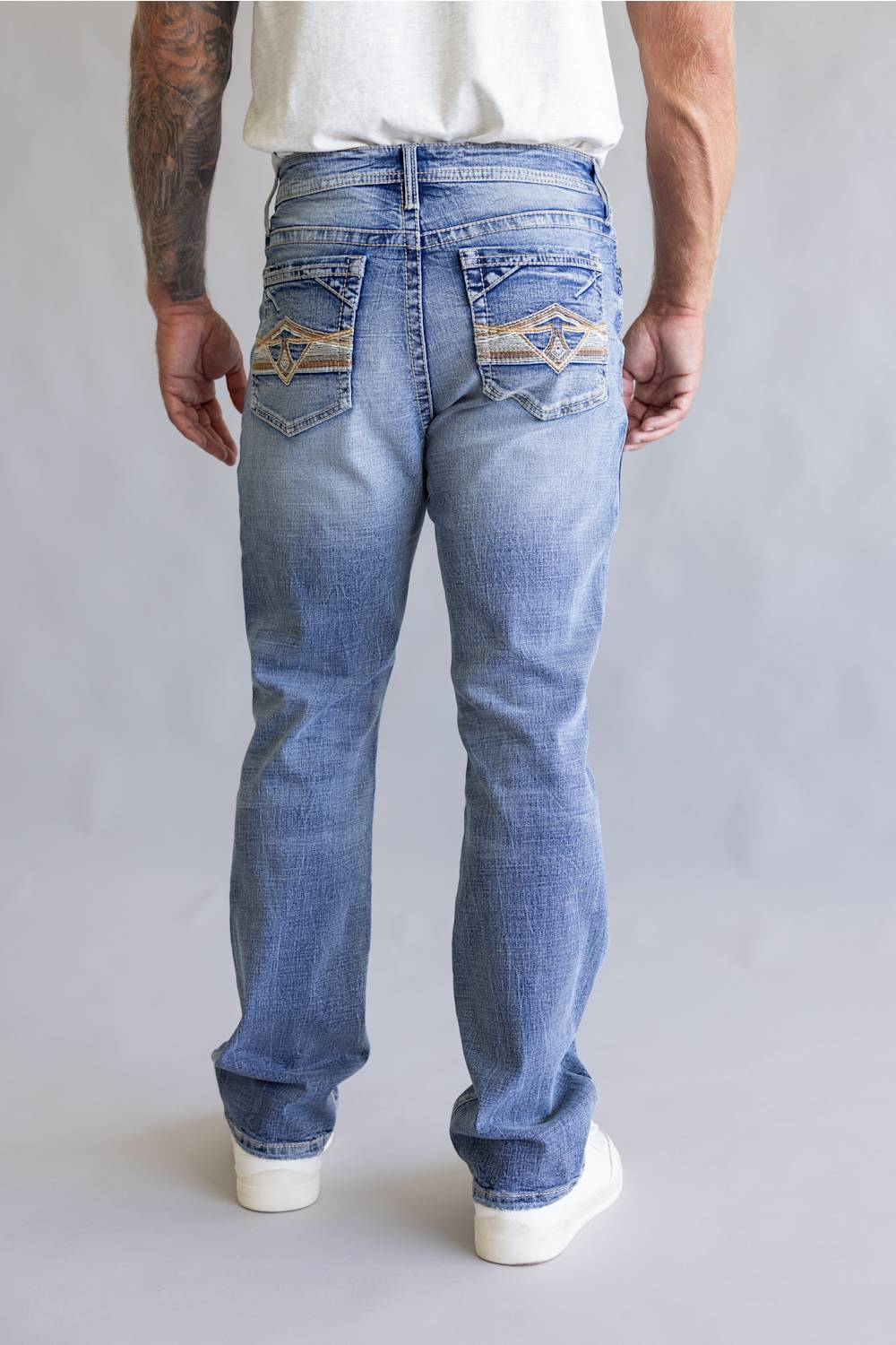 Rear view of a person wearing light blue denim jeans with decorative embroidered patch detailing on the back pockets, paired with a white t-shirt against a neutral gray background.